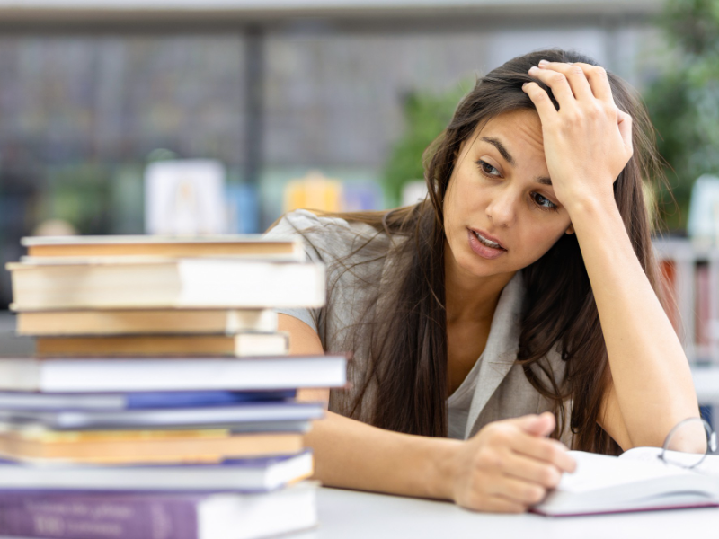 A student resting her head on her hand, looking exhausted beside a tall stack of books