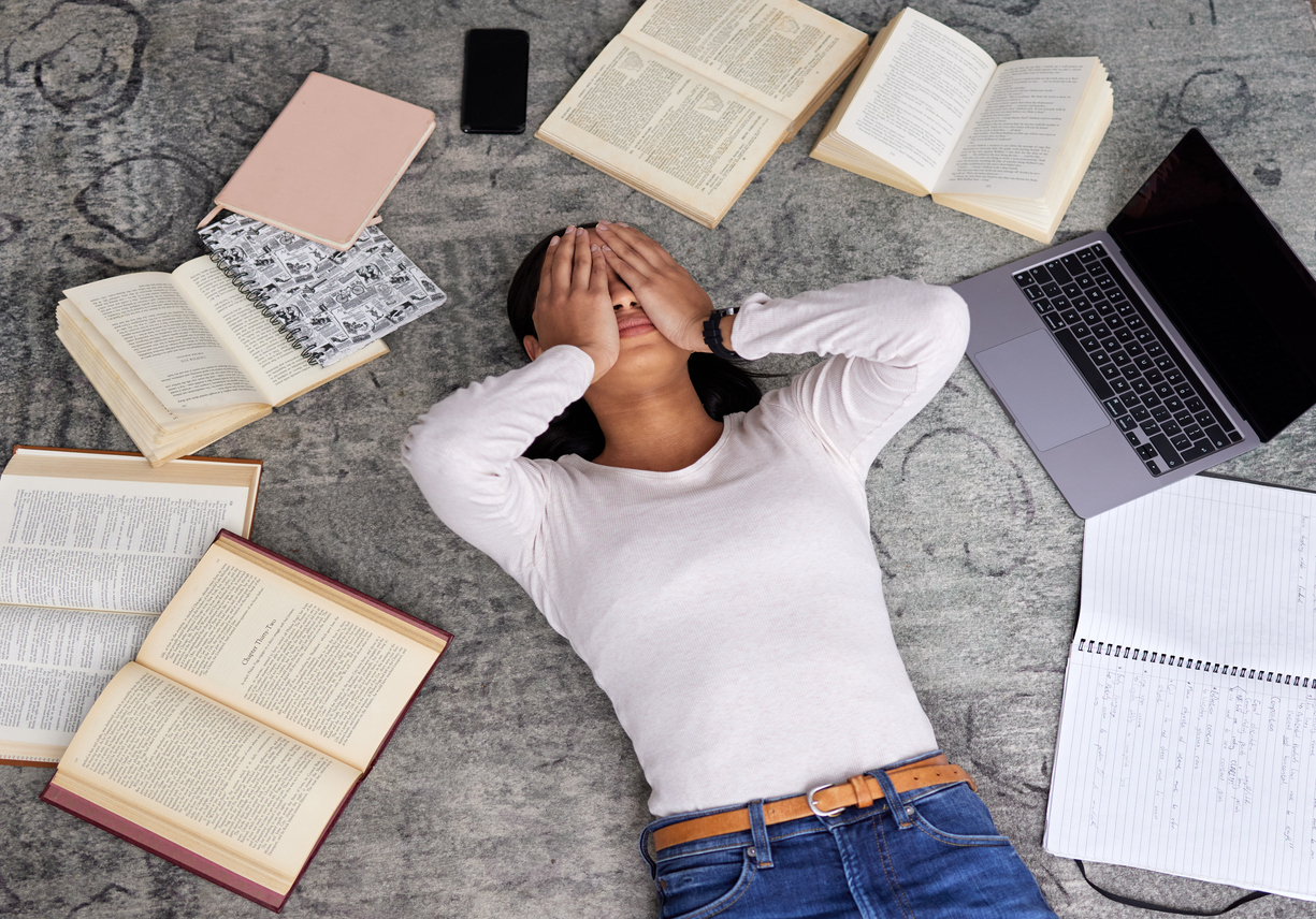 A student lying on the floor covering her eyes, overwhelmed and surrounded by books, a laptop, and notes