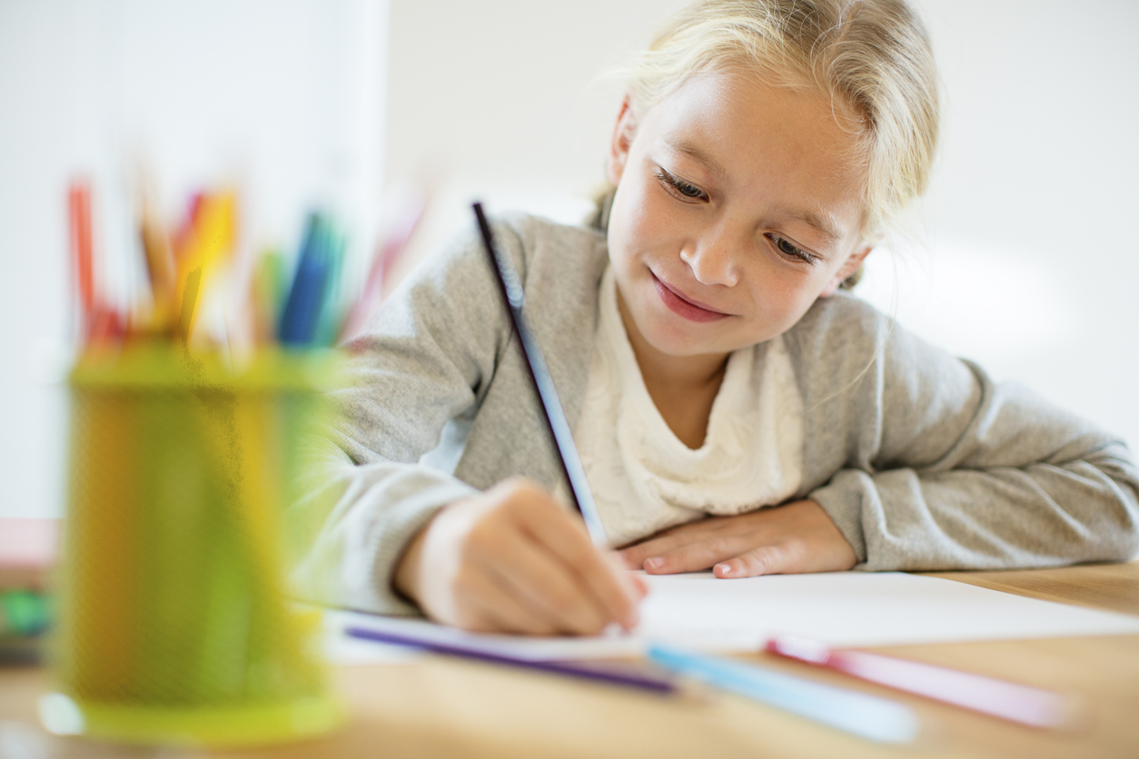 A smiling young student writing at a desk with a cup of colorful pencils nearby